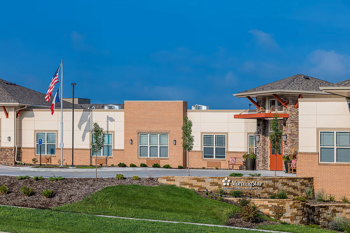 Front exterior of the MorningStar Assisted Living & Memory Care building with flags, entrance, and landscaped grounds.