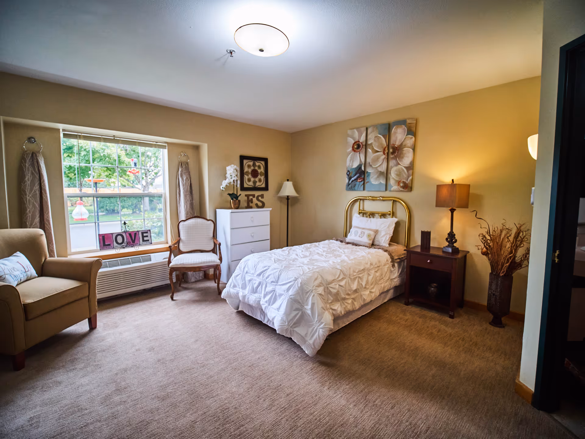 A cozy senior living bedroom with a single bed featuring a white comforter and brass headboard. The room has beige walls and carpet, a window with curtains, a beige armchair with a pillow, a wooden chair, a white dresser with decorative items, a floor lamp, a nightstand with a table lamp, and a vase with dried branches. Floral artwork hangs above the bed.