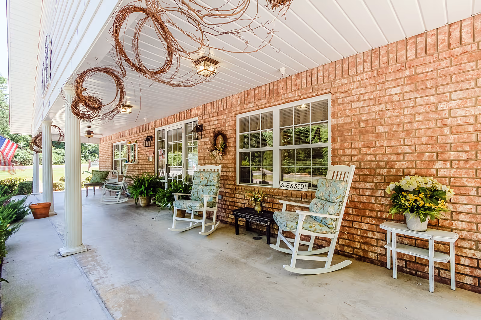 Covered porch area of Alpine House with white rocking chairs featuring floral cushions, a small black table with a flower vase, and a white side table with a flower pot. The brick wall has a window with blinds and a sign that reads 'BLESSED!'. Decorative wreaths hang on the wall and ceiling, and there are potted plants along the porch. An American flag is visible in the background near the edge of the porch.