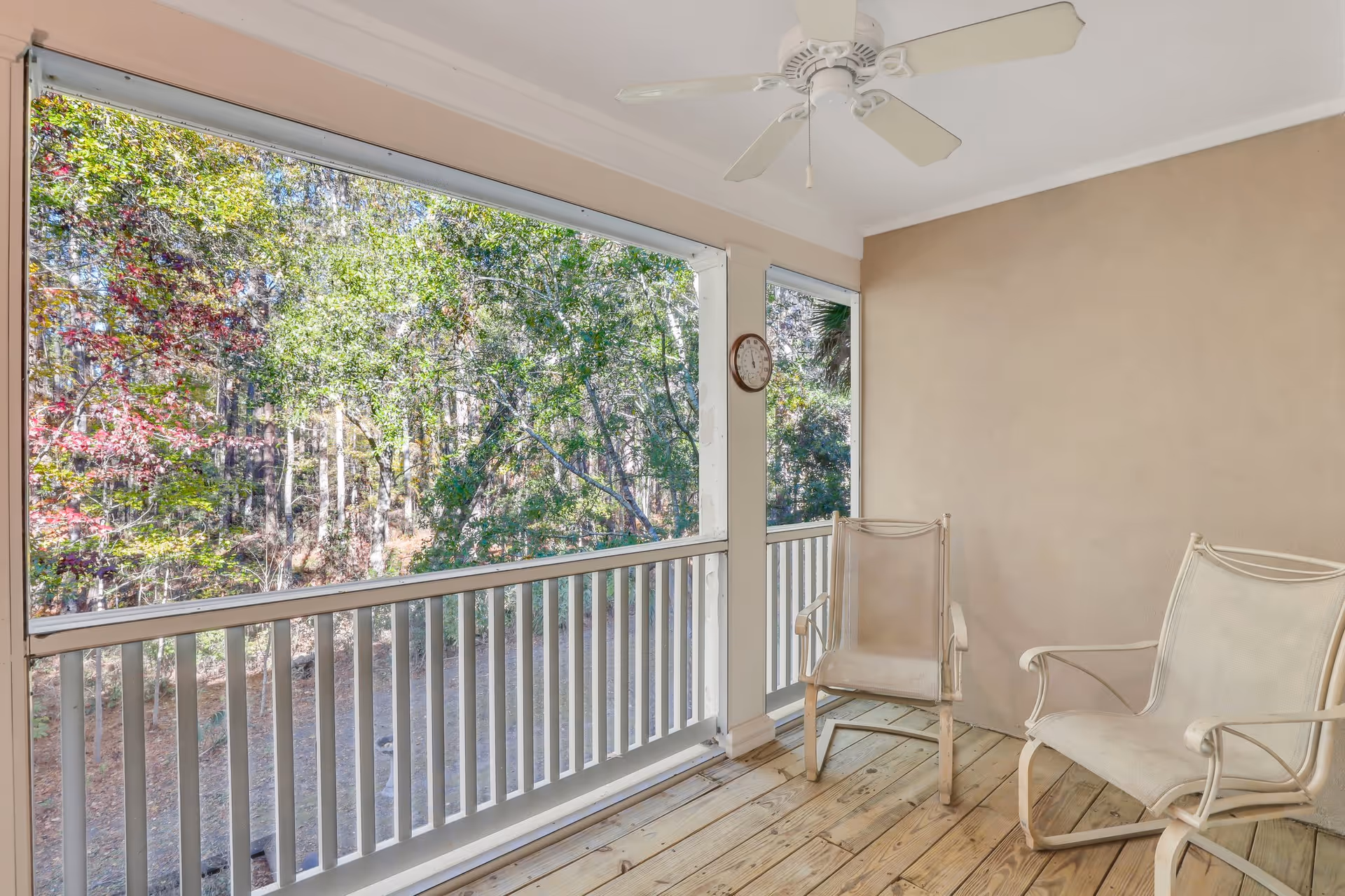 Covered screened porch with two chairs, a ceiling fan, and a railing overlooking a wooded area.