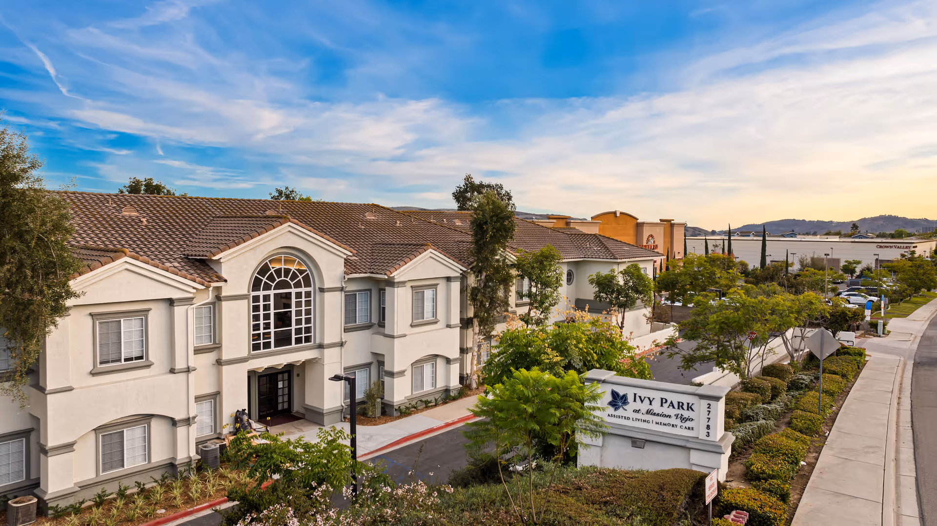 Front exterior of Ivy Park at Mission Viejo assisted living building with entrance, landscaping, and a visible sign.