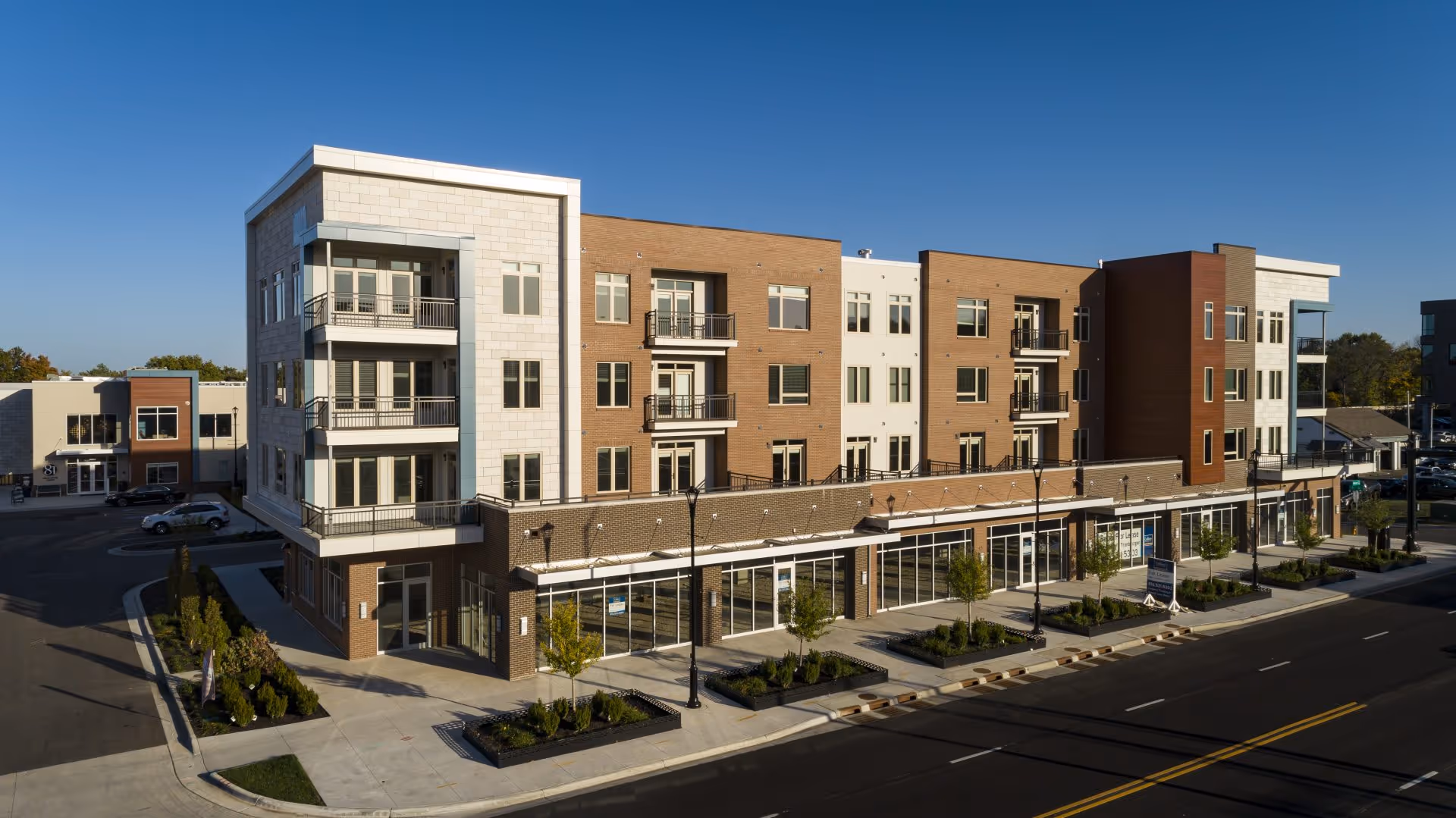 Exterior view of a modern four-story assisted and independent living facility named Avenue 81 Assisted & Independent Living, featuring multiple balconies, large windows, and a ground floor with glass storefronts. The building is surrounded by sidewalks, small landscaped areas with trees and shrubs, and a street with marked lanes in front.