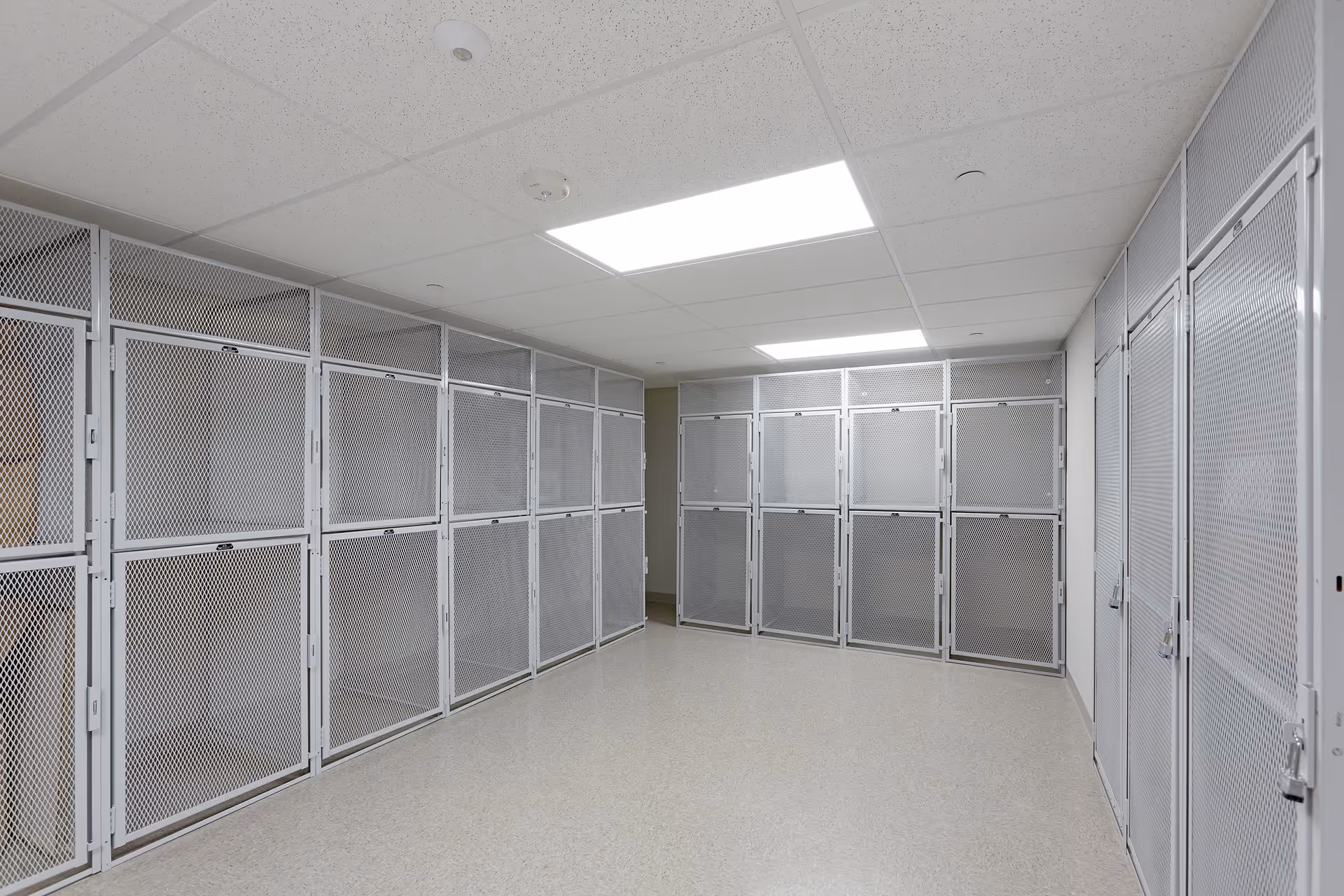 A room with multiple white metal mesh storage lockers lining the walls, illuminated by overhead fluorescent lights and featuring a light-colored floor and drop ceiling.
