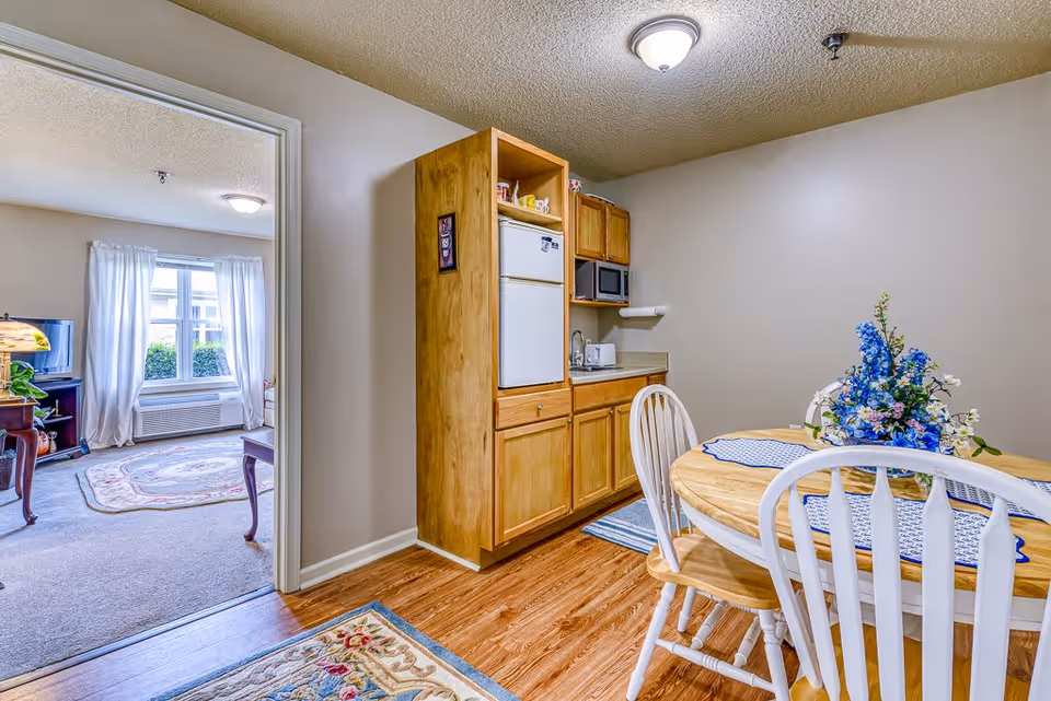 A small dining area with a round wooden table and four white chairs. The table is decorated with a blue and white table runner and a floral centerpiece with blue and white flowers. To the right, there is a kitchenette with wooden cabinets, a small refrigerator, microwave, toaster, and a sink. The floor is wood laminate, and the walls are painted beige. Through an open doorway, a living room with a carpet, window with white curtains, and a TV on a stand is visible.