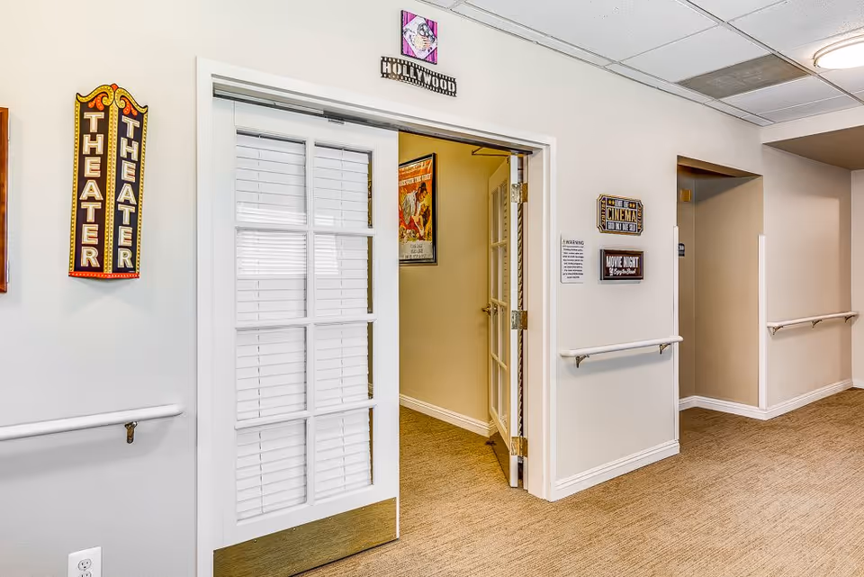 Interior hallway of a senior living facility with beige walls and carpeted floor. A white door with glass panels and blinds is open, leading to a room labeled as a theater with decorative signs on the walls including 'THEATER', 'HOLLYWOOD', 'CINEMA', and 'MOVIE NIGHT'. Handrails run along the walls of the hallway.