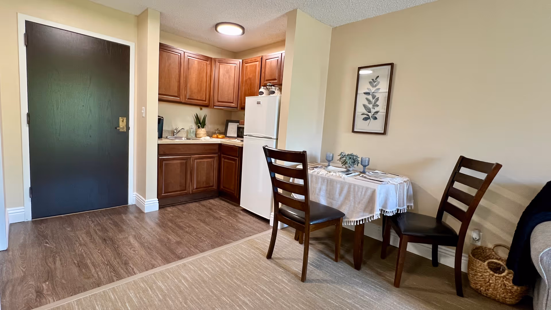 Small apartment kitchenette and dining area with wooden cabinets, a white refrigerator, and a table set for two near the entrance door.
