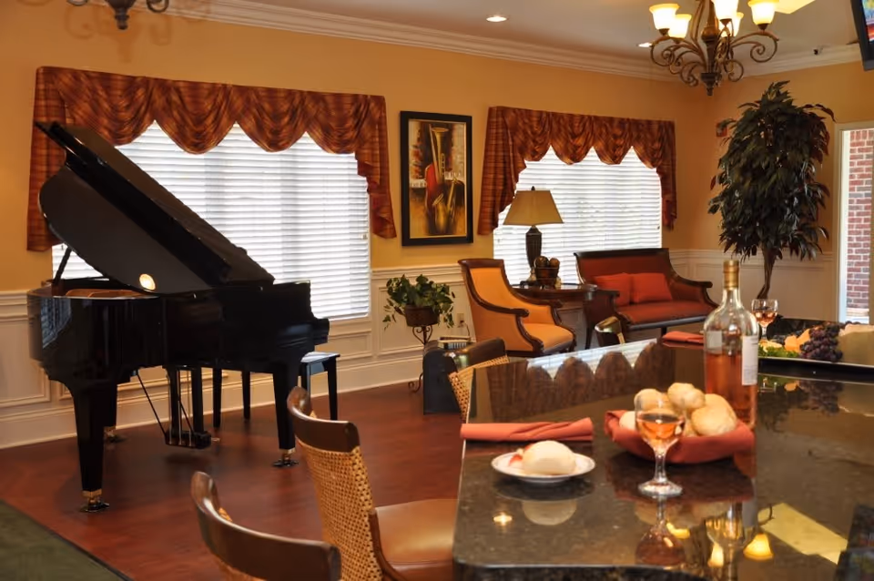 A cozy interior room featuring a black grand piano near two large windows with red valance curtains. The room has wooden flooring, a seating area with a red cushioned bench and two armchairs, a side table with a lamp, and a framed painting on the wall. In the foreground, there is a polished countertop with a bottle of wine, a glass of rosé, a plate with bread rolls, and folded red napkins. A chandelier hangs from the ceiling, and a tall potted plant is near the doorway.