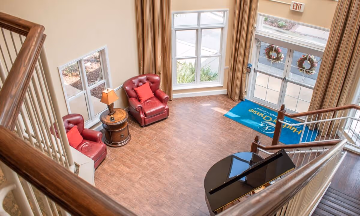 View from the staircase overlooking a lobby area with two red leather armchairs with red cushions, a round wooden side table with a lamp, large windows with beige curtains, a glass entrance door decorated with wreaths, and a black grand piano in the corner.