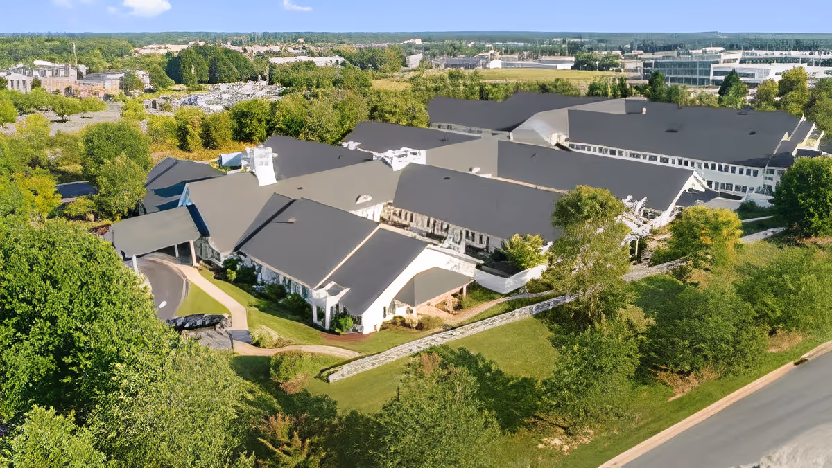 Aerial view of a large assisted living facility with dark roofs surrounded by trees and landscaped grounds.