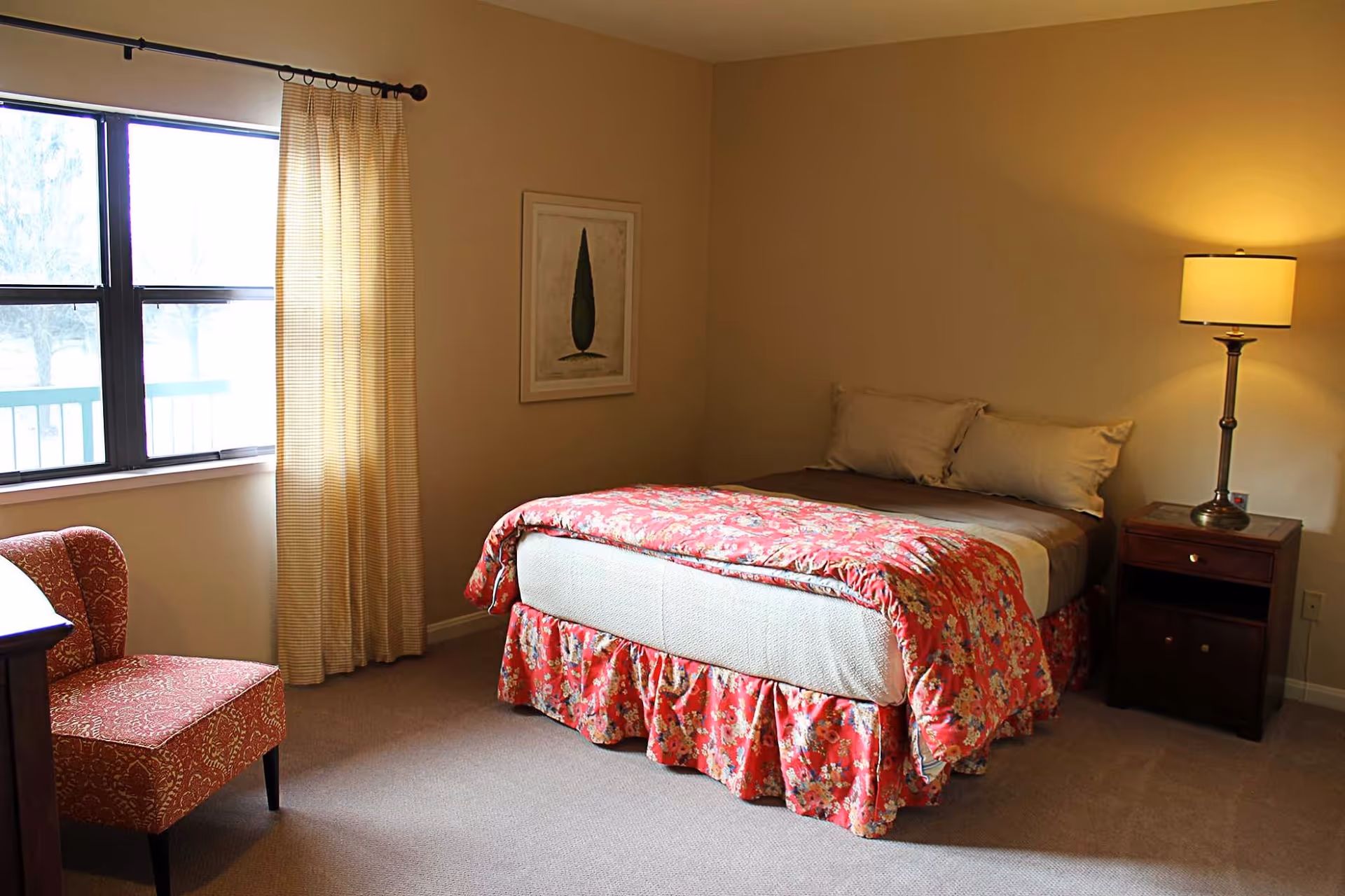 A neatly furnished bedroom with a bed in a floral bedspread, a nightstand and lamp, a window with curtains, and an upholstered chair.