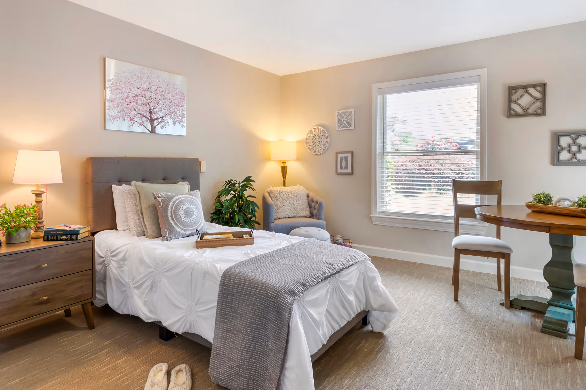 Bright, cozy bedroom with a made bed, bedside tables and lamps, an armchair, a window with blinds, and a small table and chairs.