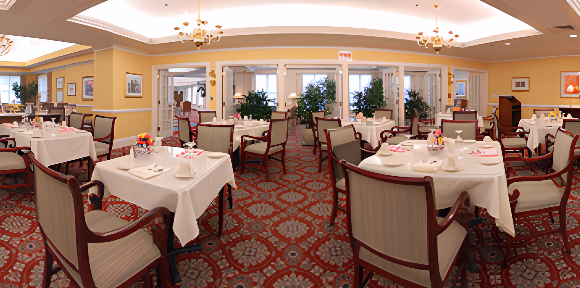 Bright, formal dining room with multiple tables set with white tablecloths and chairs on a patterned carpet.
