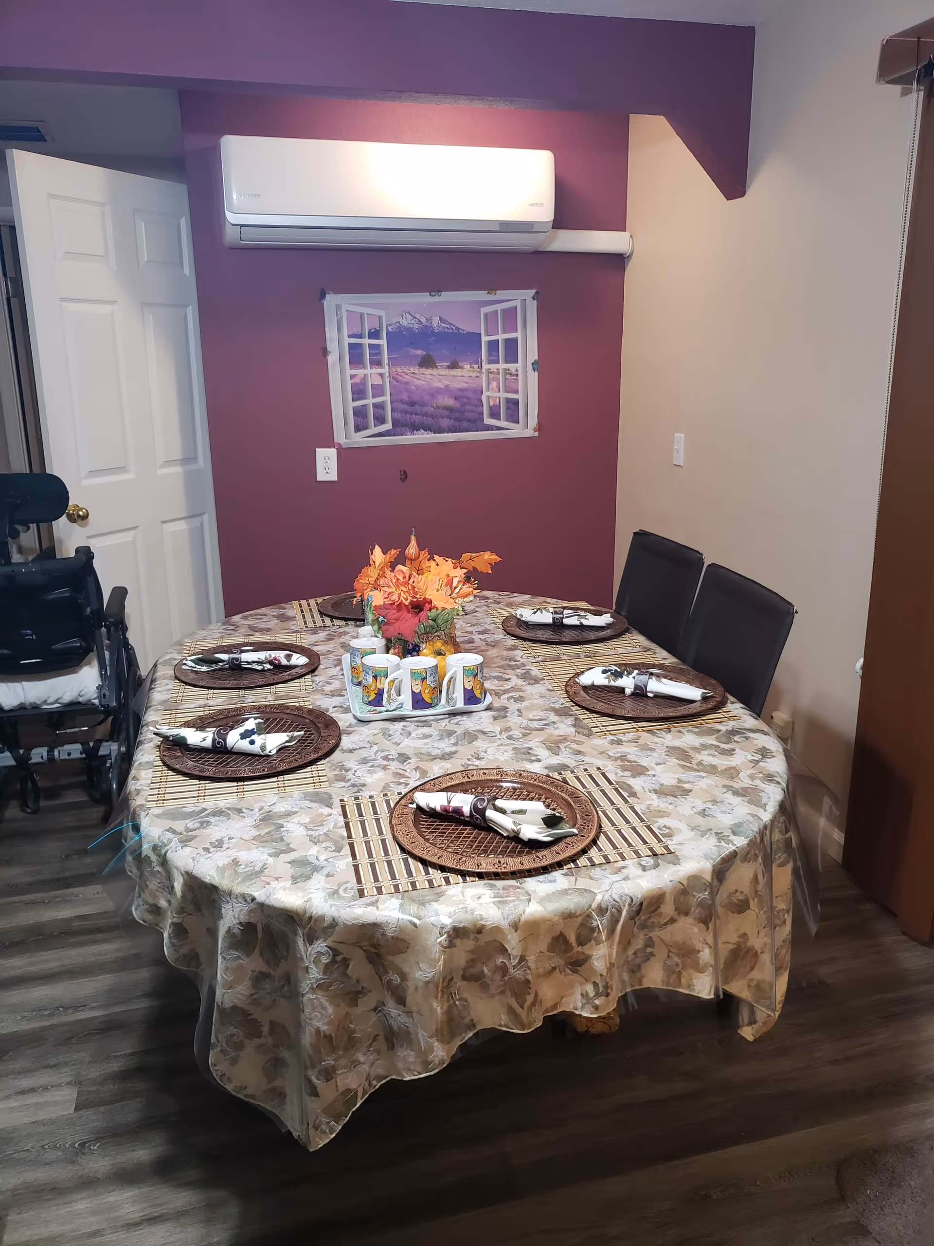 Dining room with a round table set for six with placemats and a floral tablecloth, a centerpiece, chairs and a wheelchair near a purple accent wall with an air conditioner and framed landscape art.