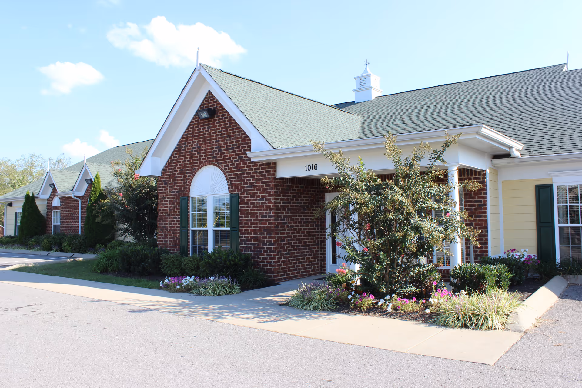 Exterior view of a senior living facility building with red brick and yellow siding, green shutters, and a green roof. The entrance is surrounded by well-maintained landscaping with bushes and colorful flowers. The building number 1016 is visible above the entrance.