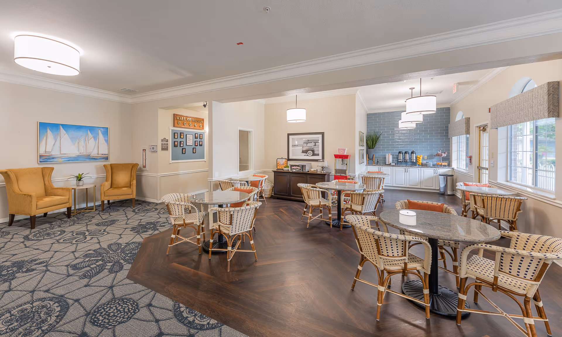 A bright and inviting common dining area with several round tables and wicker chairs. The floor has a combination of patterned carpet and wood. There are two mustard yellow armchairs with a small table between them on the left side. The back wall features a 'Wall of Honor' display and a sideboard with a popcorn machine and other snacks. The far end of the room has a counter with coffee dispensers, cups, and a blue tiled backsplash. Large windows on the right side let in natural light.