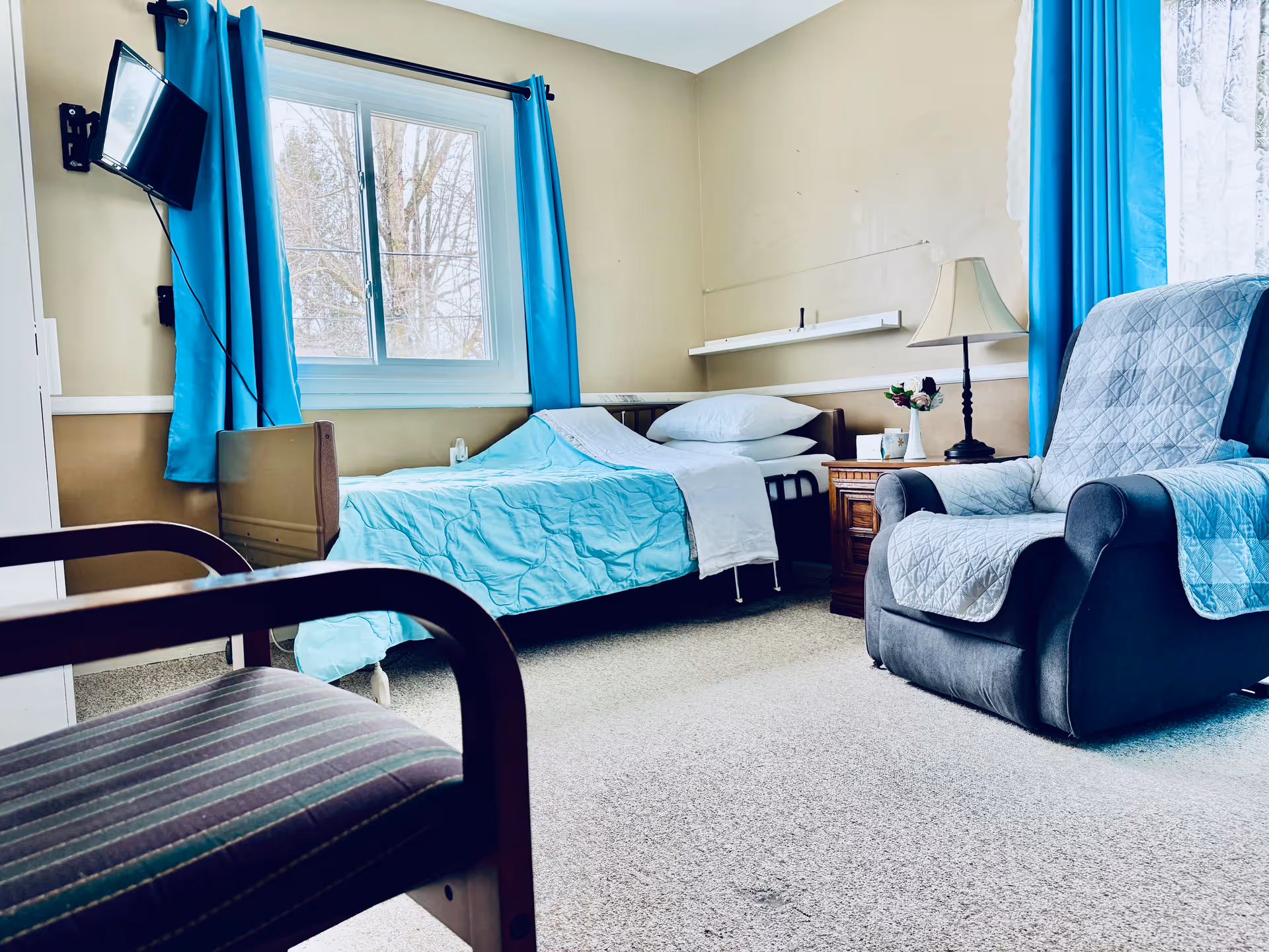 A cozy bedroom in a senior living facility with a single bed covered in a light blue blanket and white pillows. There is a window with bright blue curtains letting in natural light. Next to the bed is a wooden nightstand with a lamp and a small vase with flowers. A comfortable armchair with a quilted cover is positioned near the window, and a striped chair is partially visible in the foreground. A small wall-mounted TV is angled towards the bed.