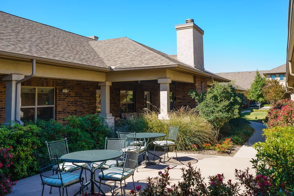 Outdoor courtyard patio with metal tables and chairs in front of a brick assisted living building surrounded by landscaping.