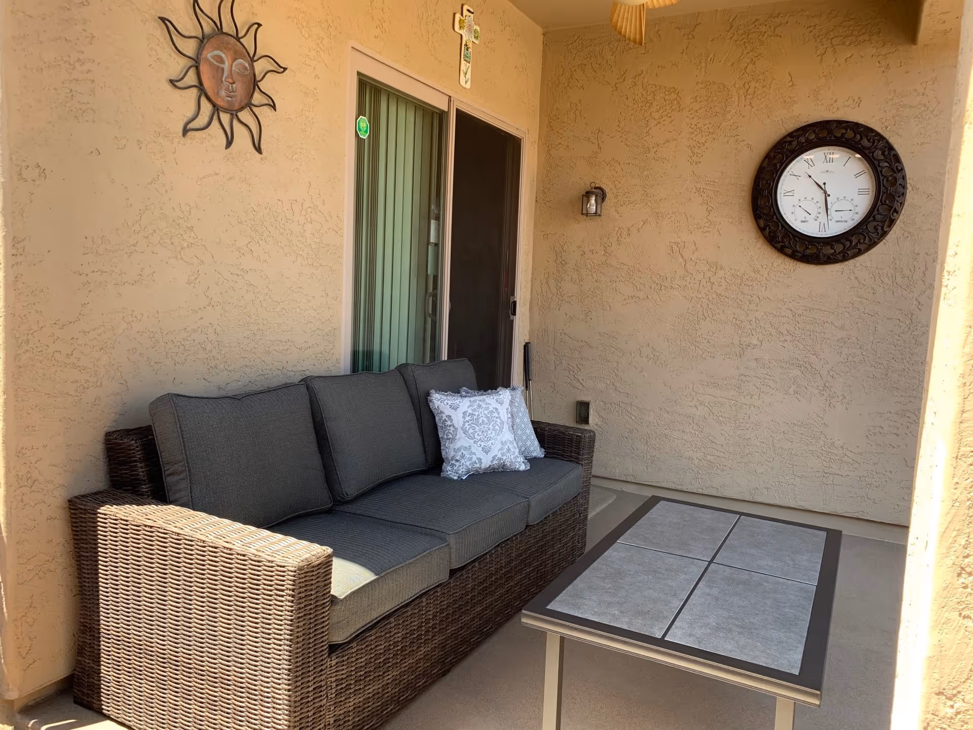 A covered outdoor patio area with a wicker sofa featuring gray cushions and a decorative pillow, a rectangular coffee table with a tiled top, a wall clock with Roman numerals, a sun-shaped wall decoration, and a small cross hanging above a sliding glass door.