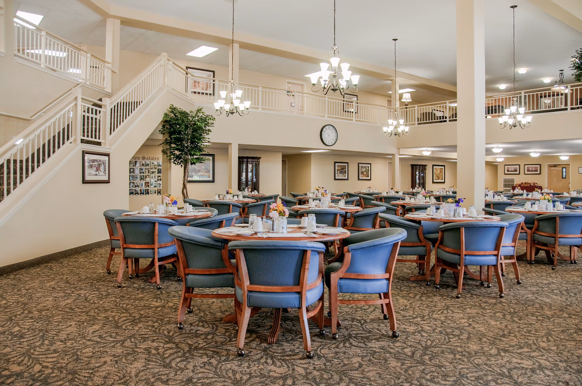Spacious senior dining room with round tables and blue upholstered chairs under chandeliers in a two-story common area.