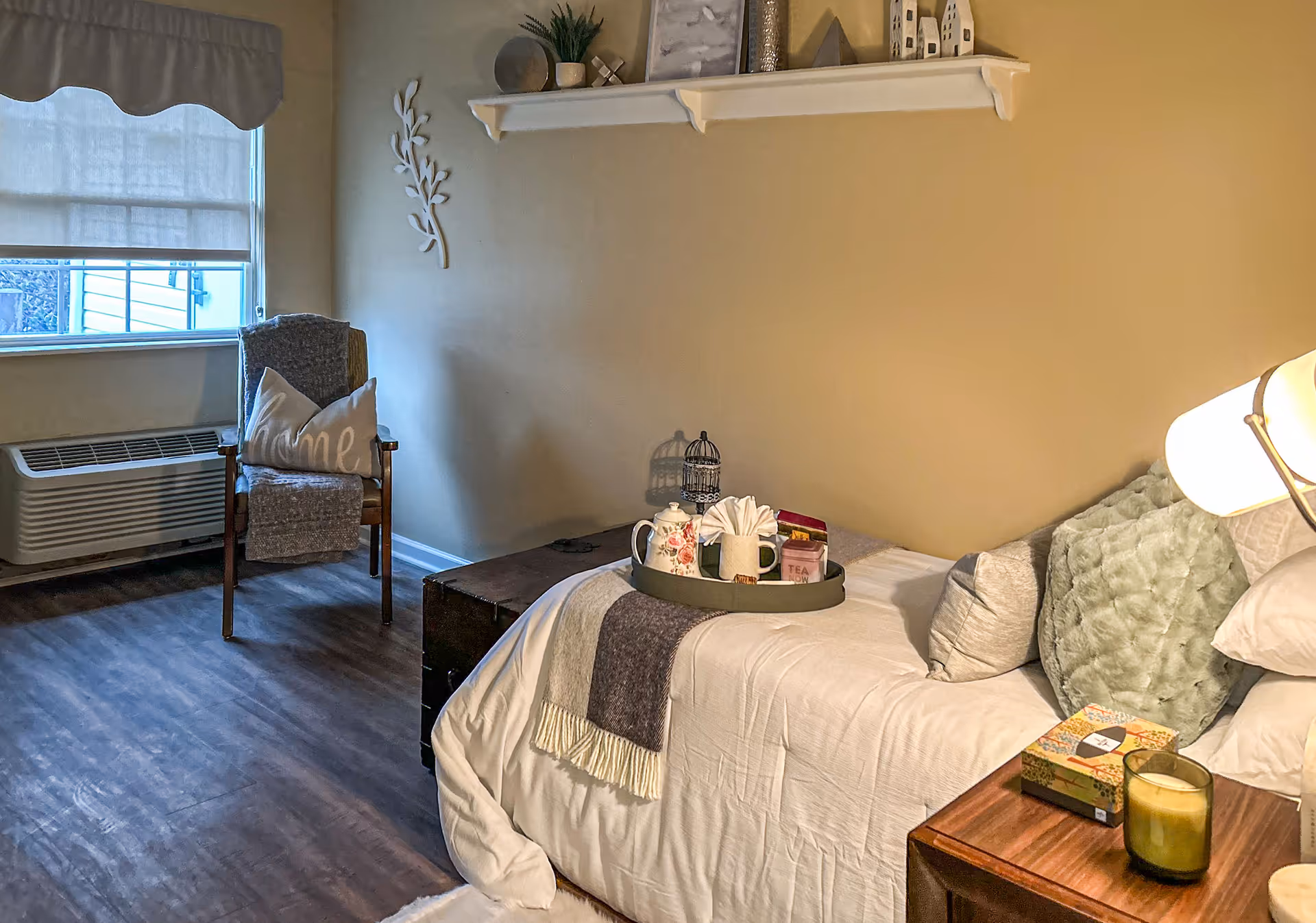 Cozy single bedroom with a made bed, bedside table and a chair by a window in a memory care facility.