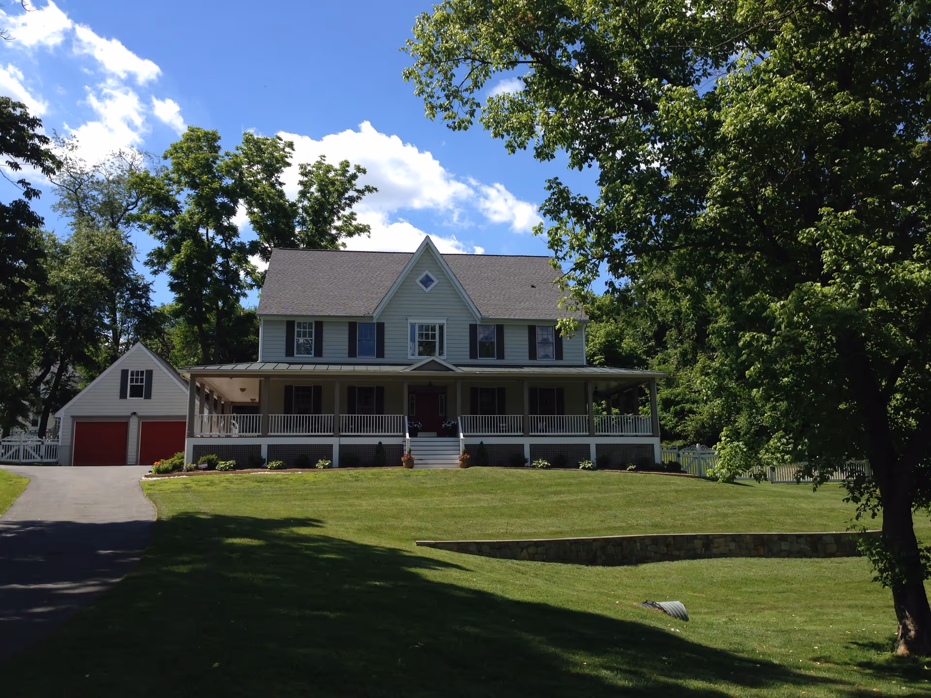 A large two-story house with a wraparound porch and a detached garage with red doors, surrounded by green grass and trees under a blue sky with some clouds.