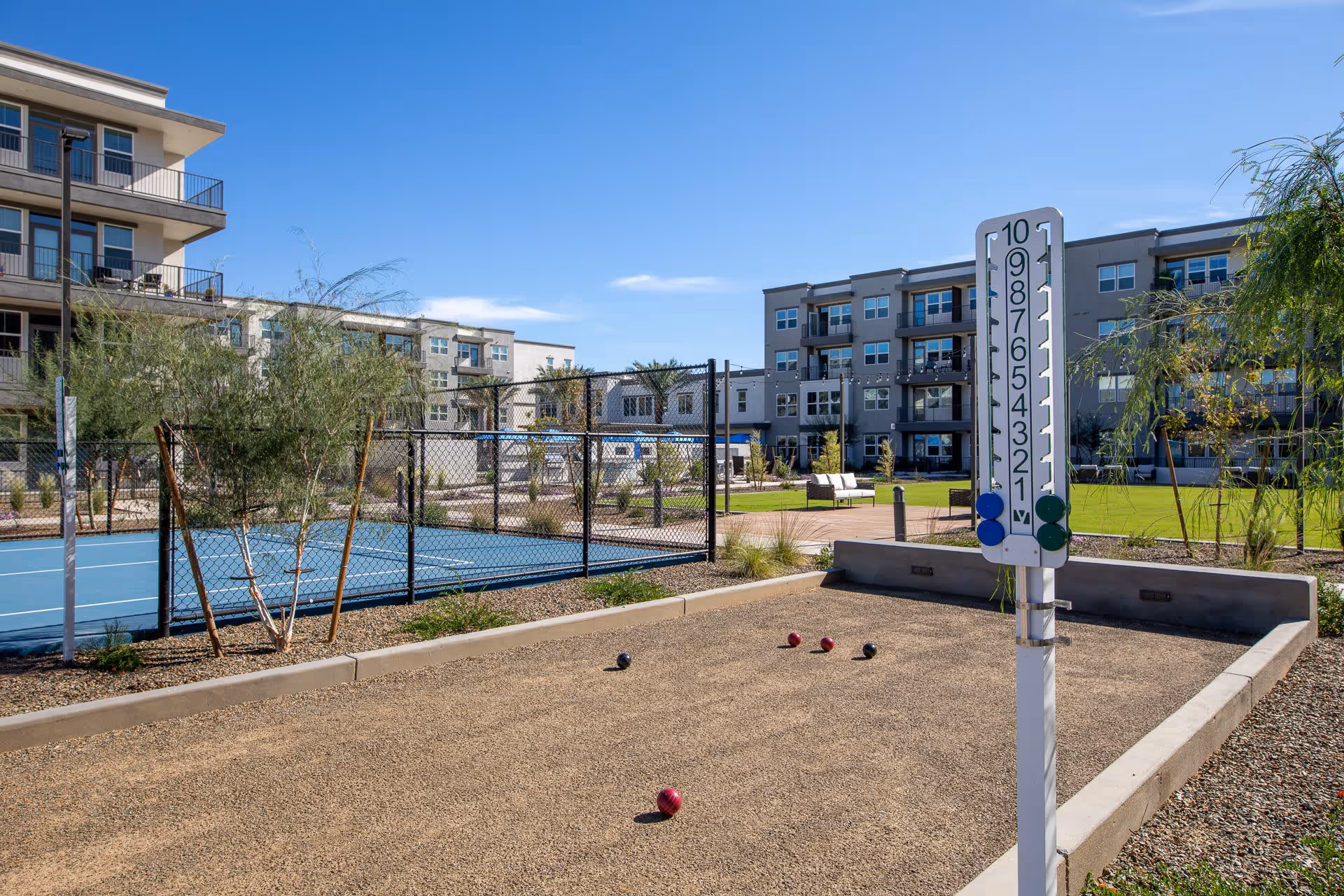 Outdoor recreational area at Mera City Center featuring a bocce ball court with balls and a scoring board, a fenced blue tennis or pickleball court, green lawn with seating, and multi-story residential buildings in the background under a clear blue sky.