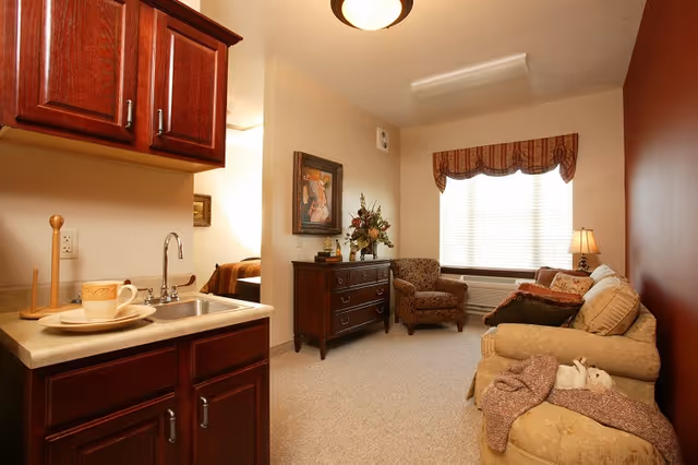 A cozy living area with a small kitchenette on the left, sofa and armchairs, a dresser with decor, and a window with a valance.