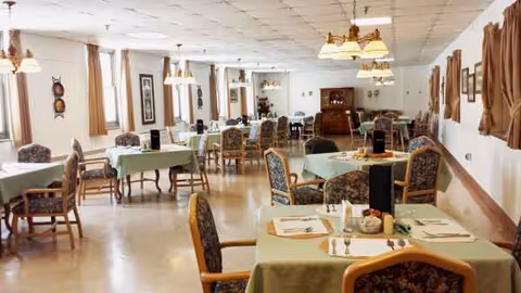 Sunlit dining room with multiple tables set for meals and cushioned chairs.