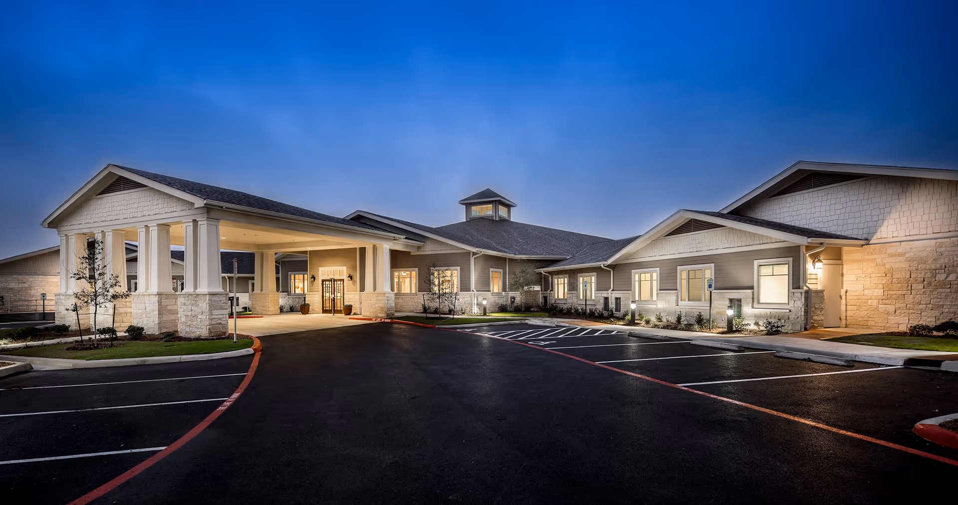 Exterior view of a single-story senior living facility building at dusk with well-lit entrance, parking spaces, and landscaped surroundings.
