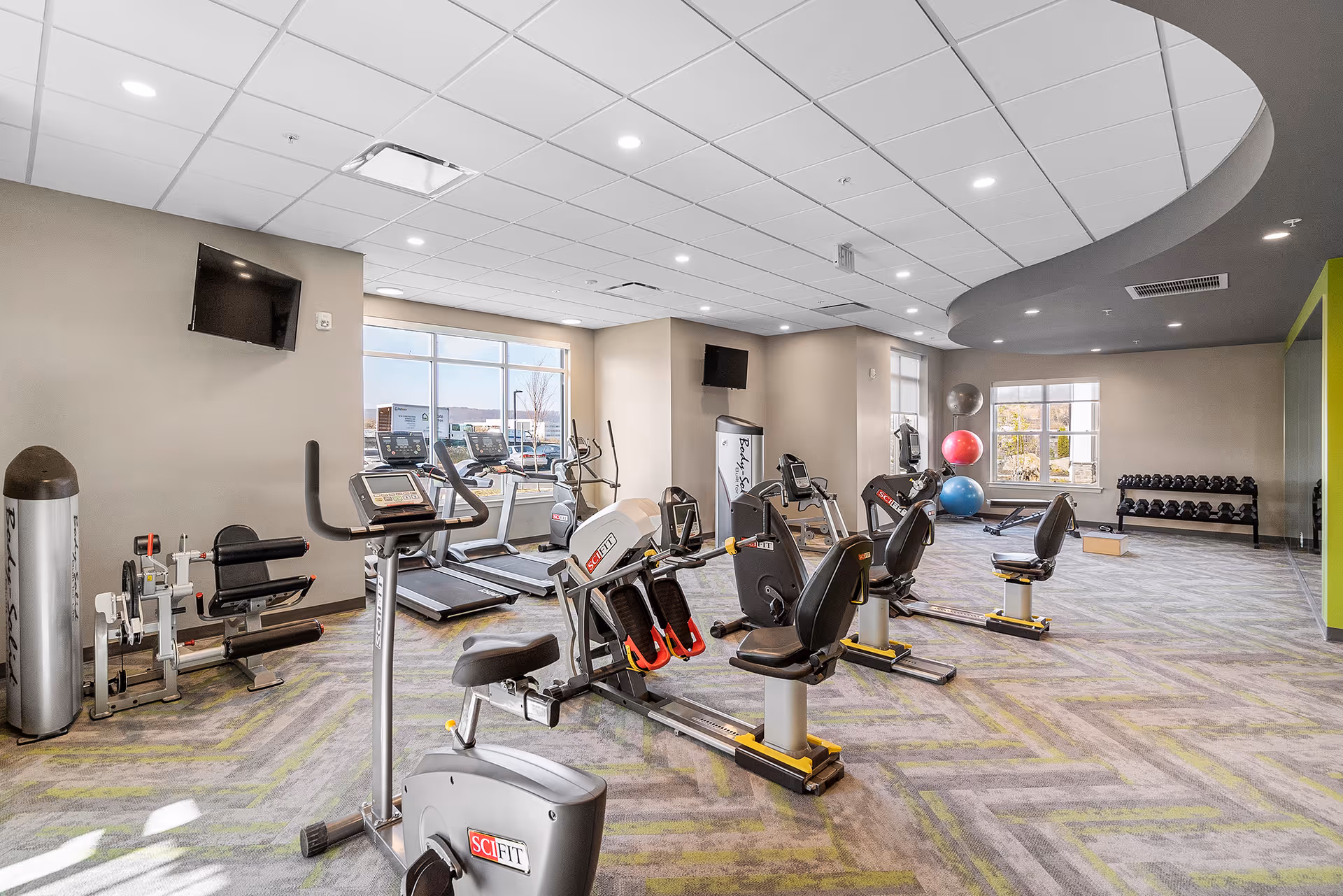 A bright and spacious fitness room with various exercise equipment including stationary bikes, treadmills, weight machines, and free weights. The room has large windows allowing natural light, a patterned carpet floor, and a ceiling with recessed lighting. There are also colorful exercise balls and a wall-mounted TV.