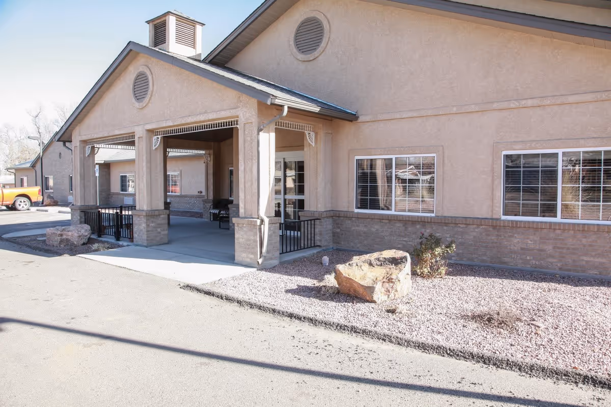 Exterior view of a beige stucco building with a covered entrance supported by columns, windows with white frames, and a gravel landscaping area with rocks and a small plant in front.