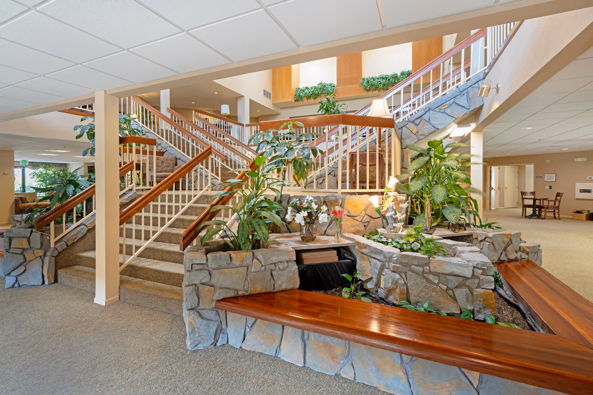 Spacious senior living facility interior with a central stone planter and indoor plants surrounded by wooden benches and a multi-level staircase.