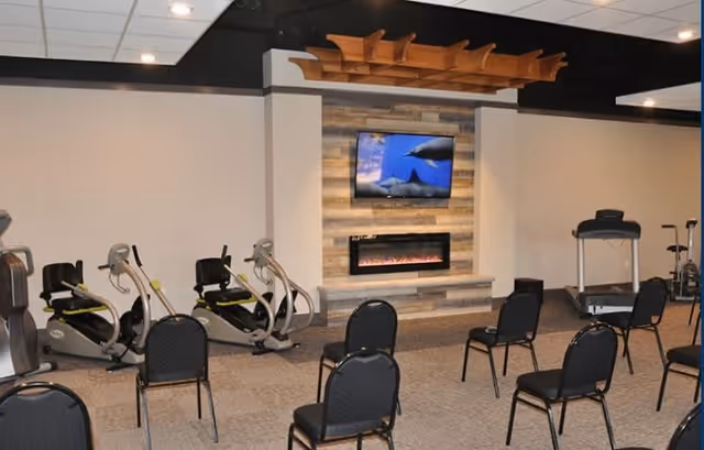 A fitness room with several exercise bikes and a treadmill arranged around a seating area with black chairs. A wall-mounted TV displays an underwater scene with dolphins above a modern electric fireplace set in a wood-paneled wall. The ceiling has recessed lighting and a decorative wooden fixture.