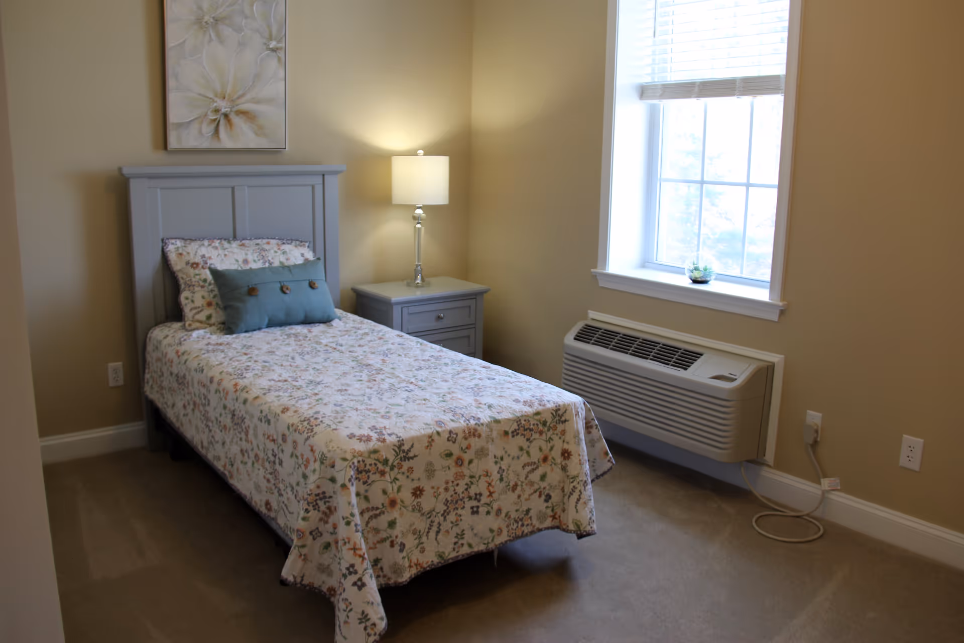 A small bedroom with a single bed covered in a floral bedspread and a decorative pillow. Next to the bed is a gray nightstand with a lamp on it. There is a window with blinds partially open, and a small plant on the windowsill. The walls are painted beige and there is a floral painting hanging above the bed.