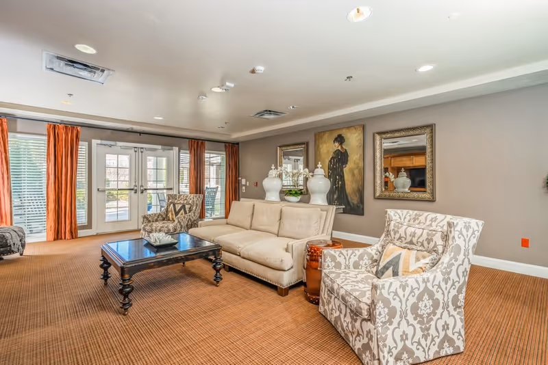 A well-lit living room area with a beige sofa, two patterned armchairs, a dark wooden coffee table, and decorative vases on a side table. The room has large windows with orange curtains, a brown carpet, and artwork on the walls including a painting of a woman and two framed mirrors.