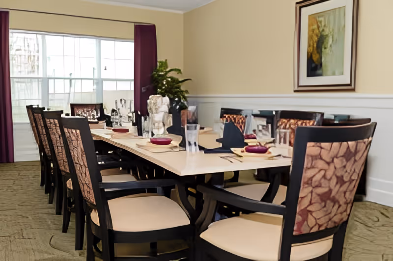 A long dining table set with plates, glasses, and napkins in a room with beige walls and a large window covered by purple curtains. The chairs around the table have patterned backs and light-colored seats. A framed painting and a potted plant are visible in the background.