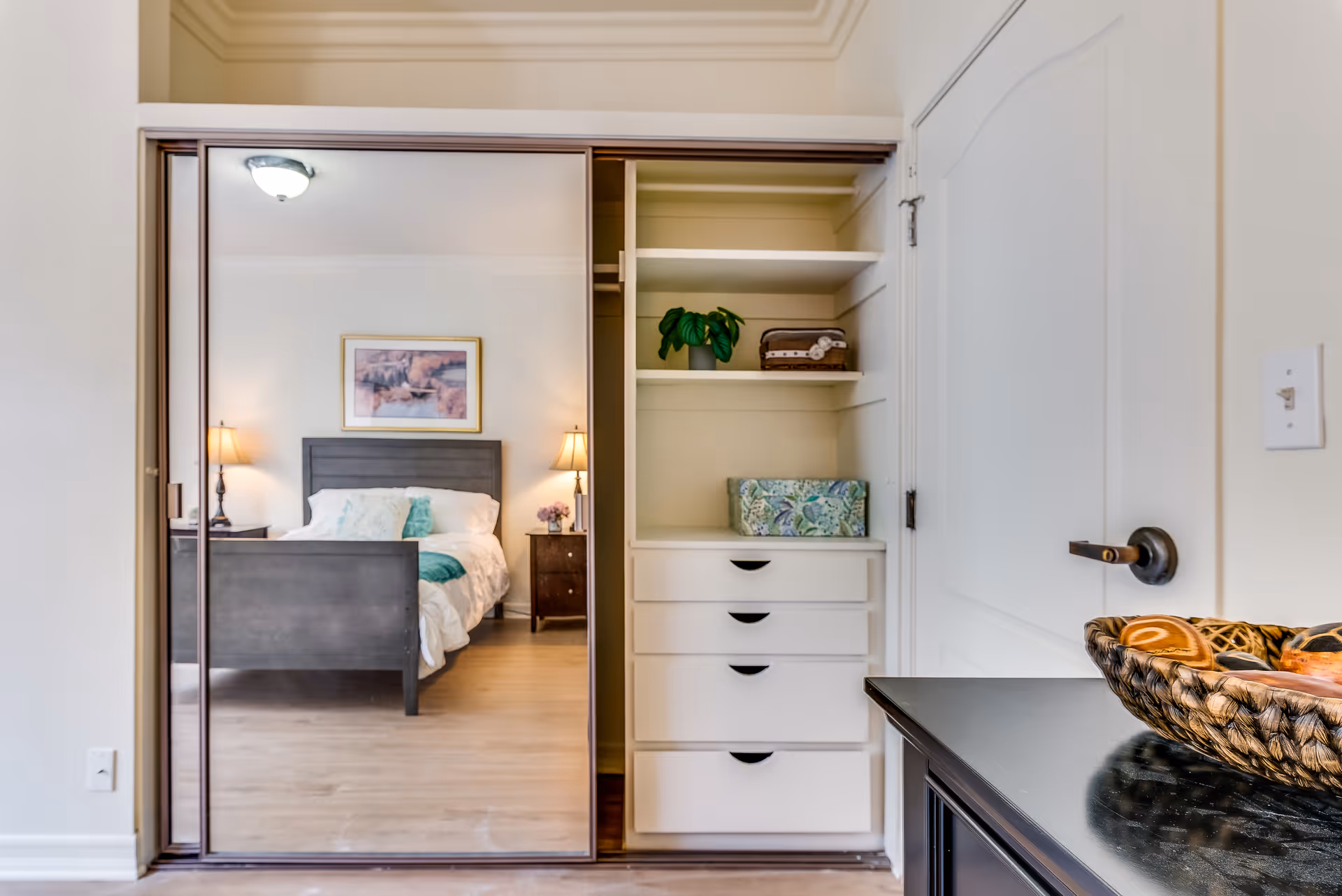 View of a bedroom reflected in a large mirrored sliding closet door. The bedroom has a dark wooden bed frame with white bedding and teal accent pillows, two bedside tables with lamps, and a framed picture on the wall. Next to the closet is a built-in shelving unit with drawers, a small plant, and decorative boxes. A dark wooden table with a woven basket is partially visible on the right side.