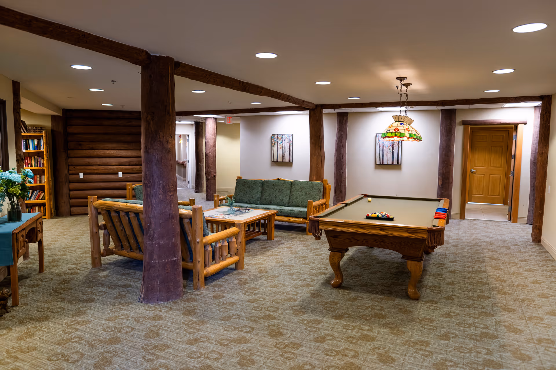 A cozy common area with rustic wooden furniture including a sofa and chairs with green cushions, a wooden coffee table, and a pool table with a stained glass hanging lamp above it. The room has carpeted floors, wooden support beams, and framed artwork on the walls. There is a bookshelf with books and flowers on a small table to the left.