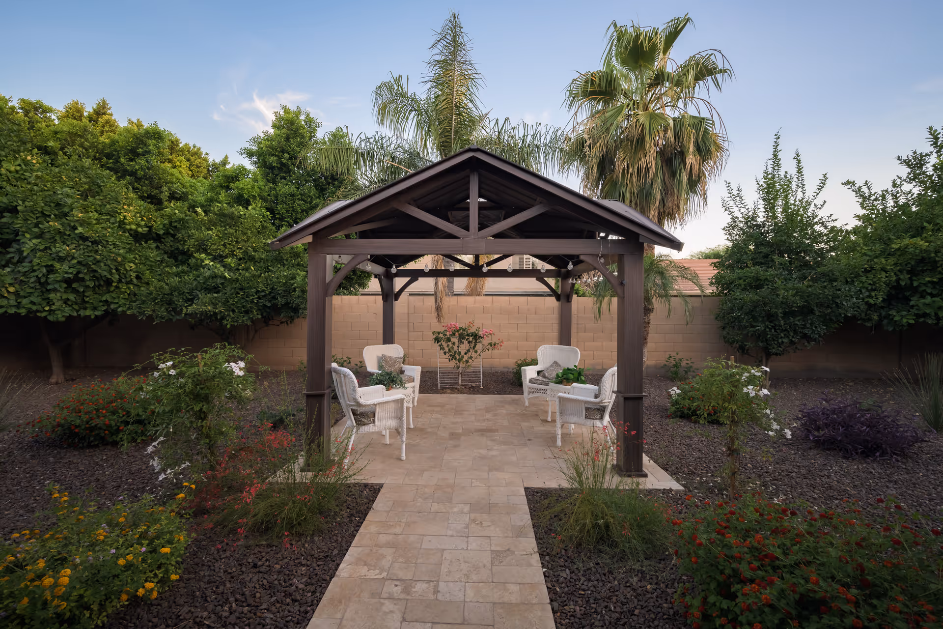 A peaceful outdoor garden area with a wooden gazebo featuring a dark brown roof and four white wicker chairs arranged inside. The gazebo is surrounded by various green plants, bushes, and trees, including palm trees, with a tiled pathway leading up to it. The sky is clear with a few wispy clouds.