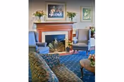 A cozy living room area with a floral patterned sofa, two blue armchairs, a wooden coffee table with a flower arrangement, and a fireplace with a decorative screen. Above the fireplace hangs a floral painting, and there are additional flower arrangements on the mantel and side table. The room has a blue carpet with a subtle pattern.