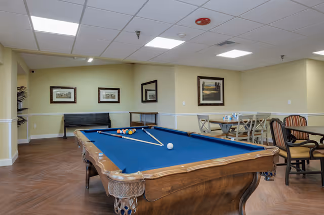 Interior view of a recreational room featuring a blue felt pool table with pool balls and cues arranged on it. The room has light yellow walls adorned with framed artwork, a wooden floor, and a ceiling with recessed lighting. In the background, there are tables and chairs for seating.