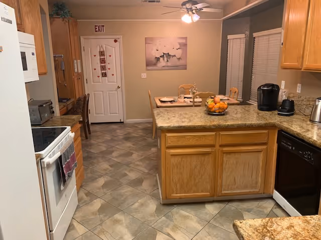 Open kitchen with a central island and wooden cabinets, stove and dishwasher visible, and a small dining table near a door under an EXIT sign.