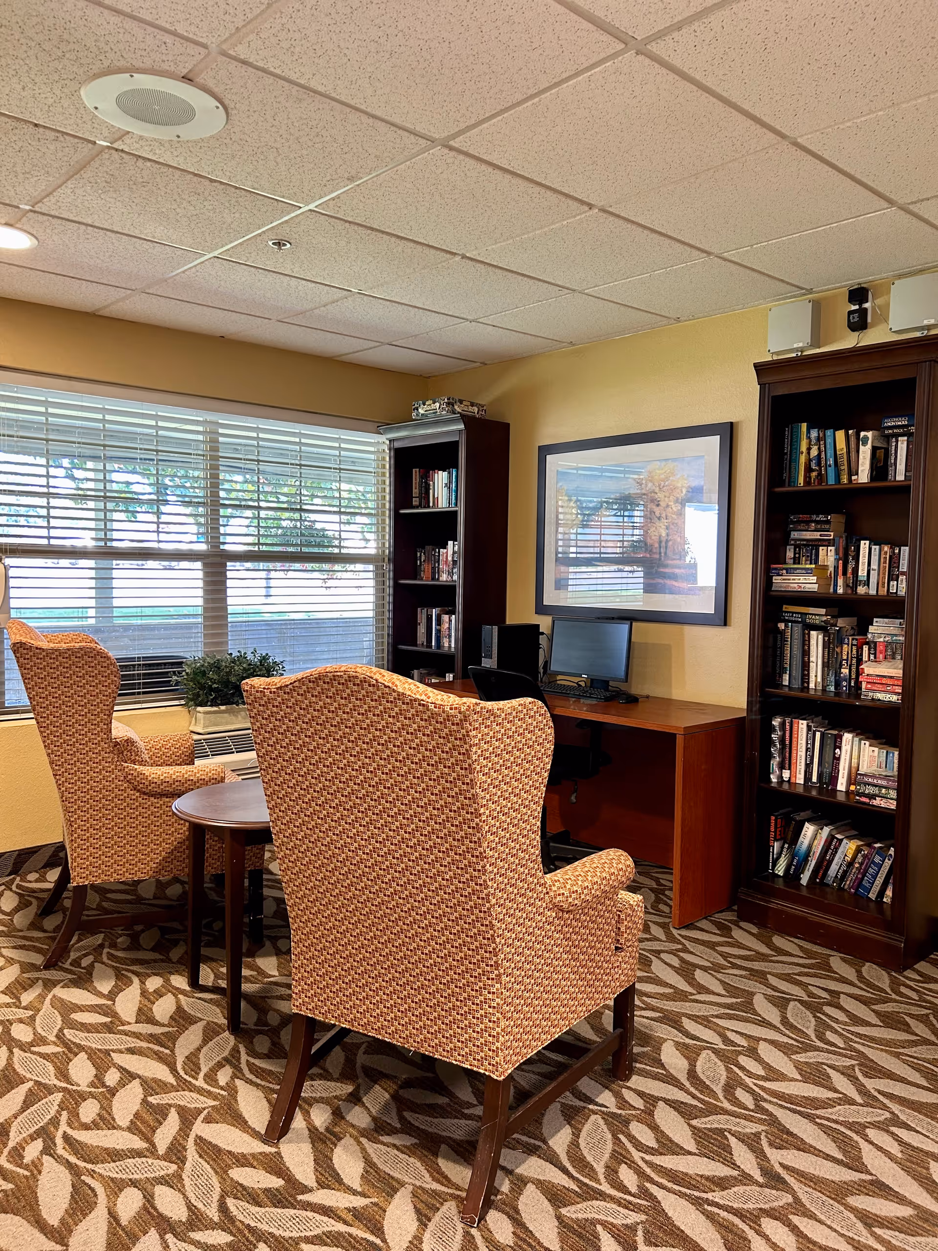 A cozy senior living common area with two patterned armchairs facing a small round wooden table. Behind the chairs is a large window with blinds partially open, letting in natural light. To the right, there is a wooden desk with a computer and a black office chair. Two tall wooden bookshelves filled with books flank the desk. A framed picture hangs on the wall above the desk. The carpet has a leaf pattern, and the walls are painted a warm beige color.