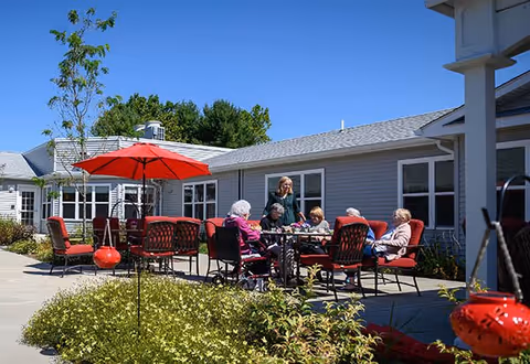 A group of elderly people sitting around a table with red cushioned chairs under a red umbrella in an outdoor patio area of a senior living facility. A caregiver stands nearby interacting with them. The building with gray siding and white trim is visible in the background under a clear blue sky.