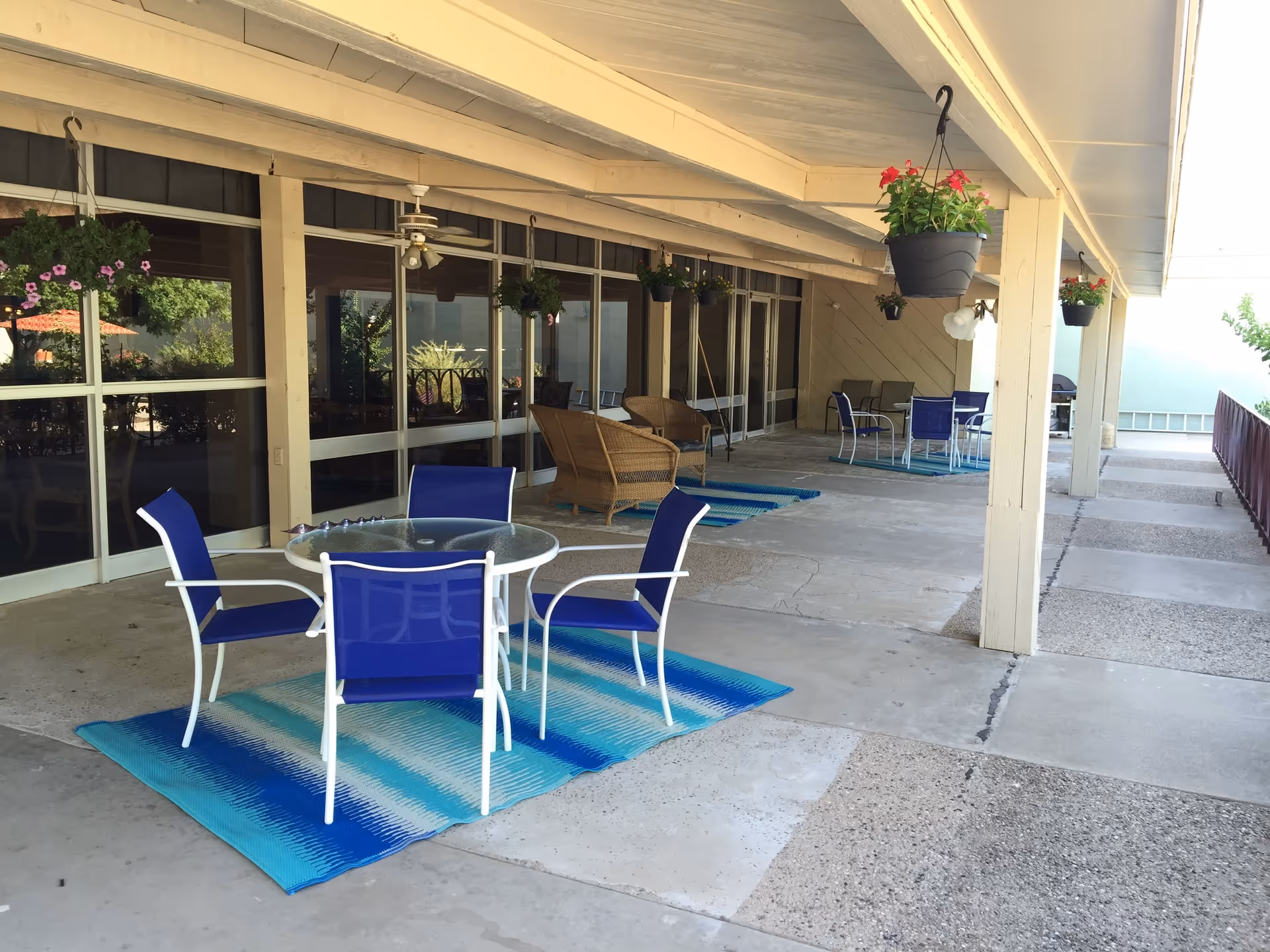 Covered outdoor patio area with multiple seating arrangements including blue chairs around glass tables and wicker chairs. Hanging flower pots with red and purple flowers are suspended from the ceiling. The patio has a concrete floor with blue and white rugs under the tables. Large windows and doors line the wall on one side.