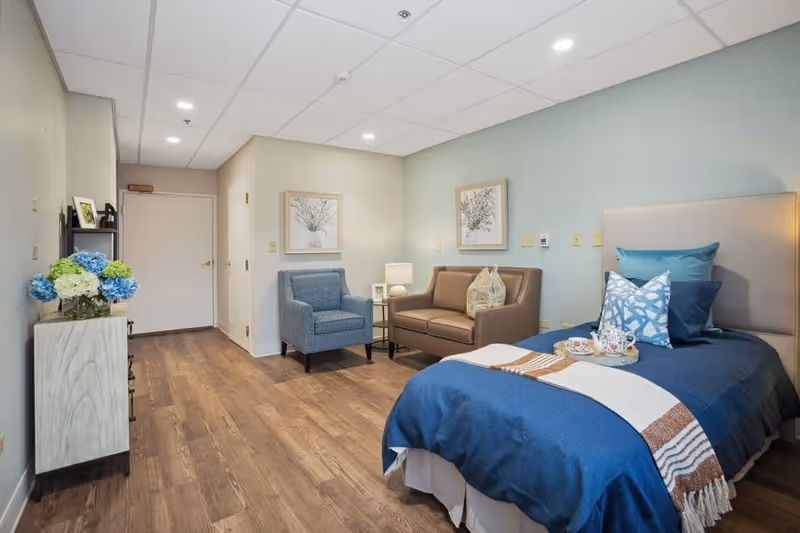 A cozy bedroom interior featuring a bed with blue bedding, a brown sofa, a blue armchair, and decorative wall art. The room has wooden flooring and a small table with tea cups.