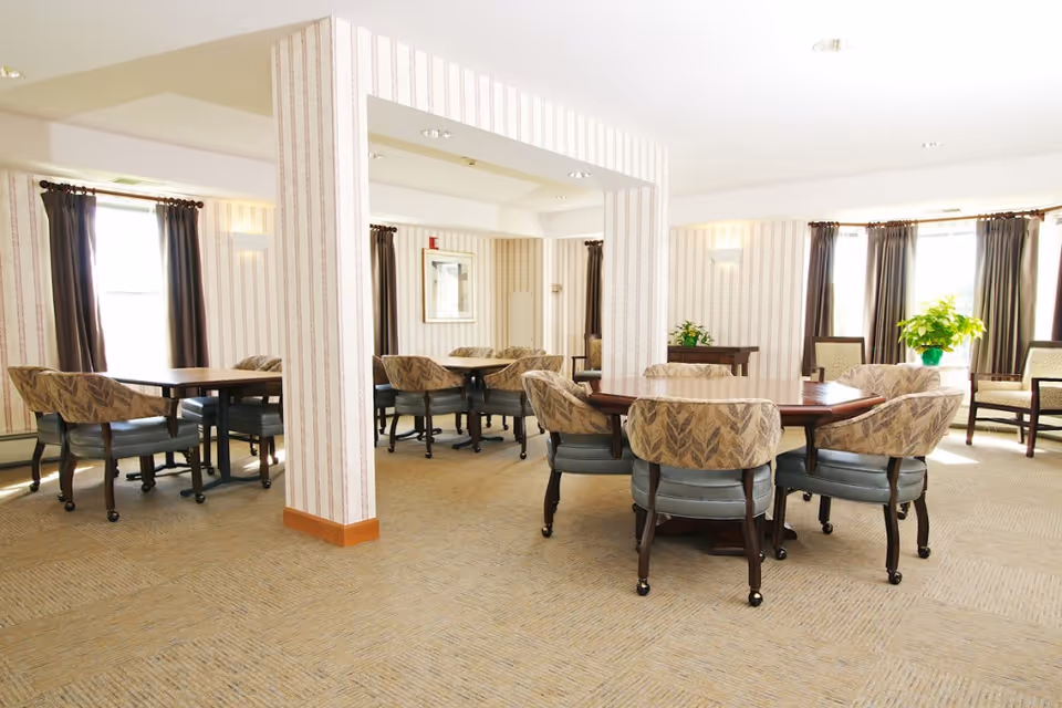 A bright and spacious dining room in a senior living facility with multiple wooden tables surrounded by cushioned chairs. The room has large windows with dark curtains allowing natural light to fill the space. The walls are decorated with light-colored striped wallpaper, and there are a few plants and framed pictures adding to the cozy atmosphere.