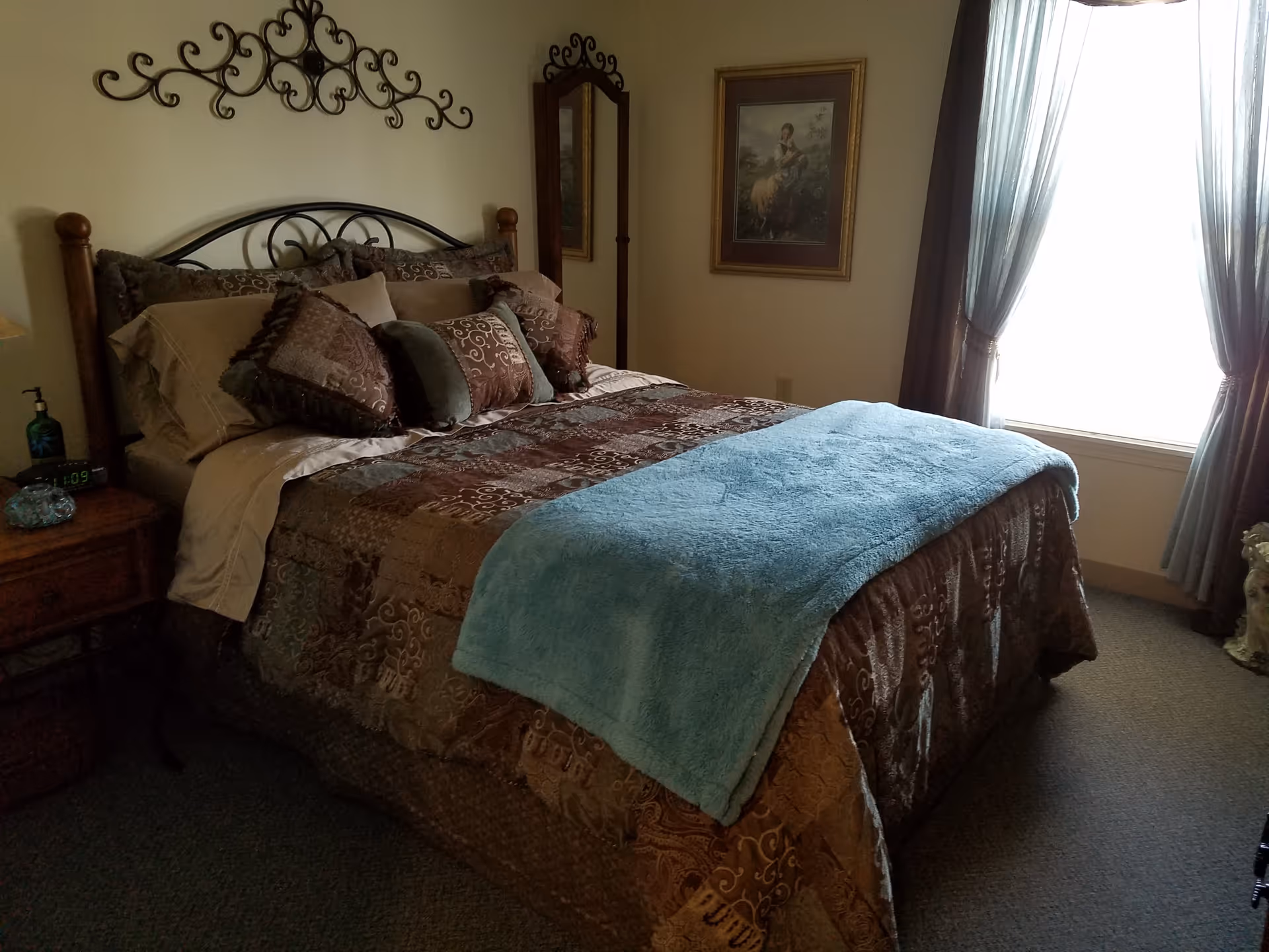 A cozy bedroom with a neatly made bed featuring a brown patterned comforter and multiple decorative pillows. A light blue throw blanket is folded at the foot of the bed. The room has a wooden nightstand with a digital clock and a bottle of hand sanitizer. There is a decorative metal wall hanging above the bed, a tall mirror in the corner, a framed painting on the wall, and a window with sheer and dark curtains allowing natural light to enter.
