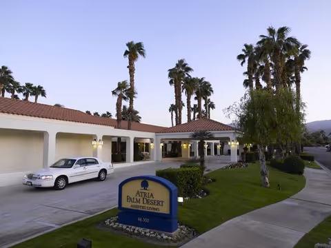 Entrance of Atria Palm Desert assisted living facility with a sign, driveway and parked car in front of a low-roof building and palm trees at dusk.