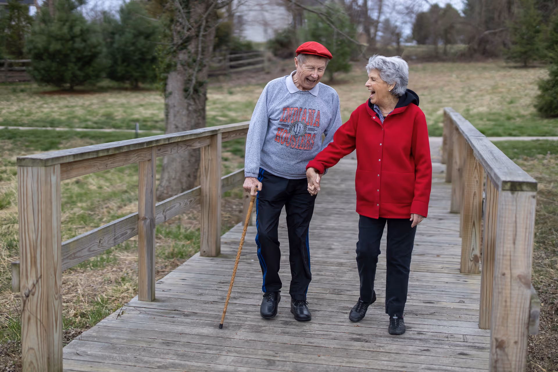 An elderly man and woman walking hand in hand on a wooden bridge outdoors. The man is using a cane and wearing a red cap and a gray Indiana Hoosiers sweatshirt. The woman is wearing a red jacket and black pants. They are smiling and appear to be enjoying their walk in a natural setting with grass and trees around.